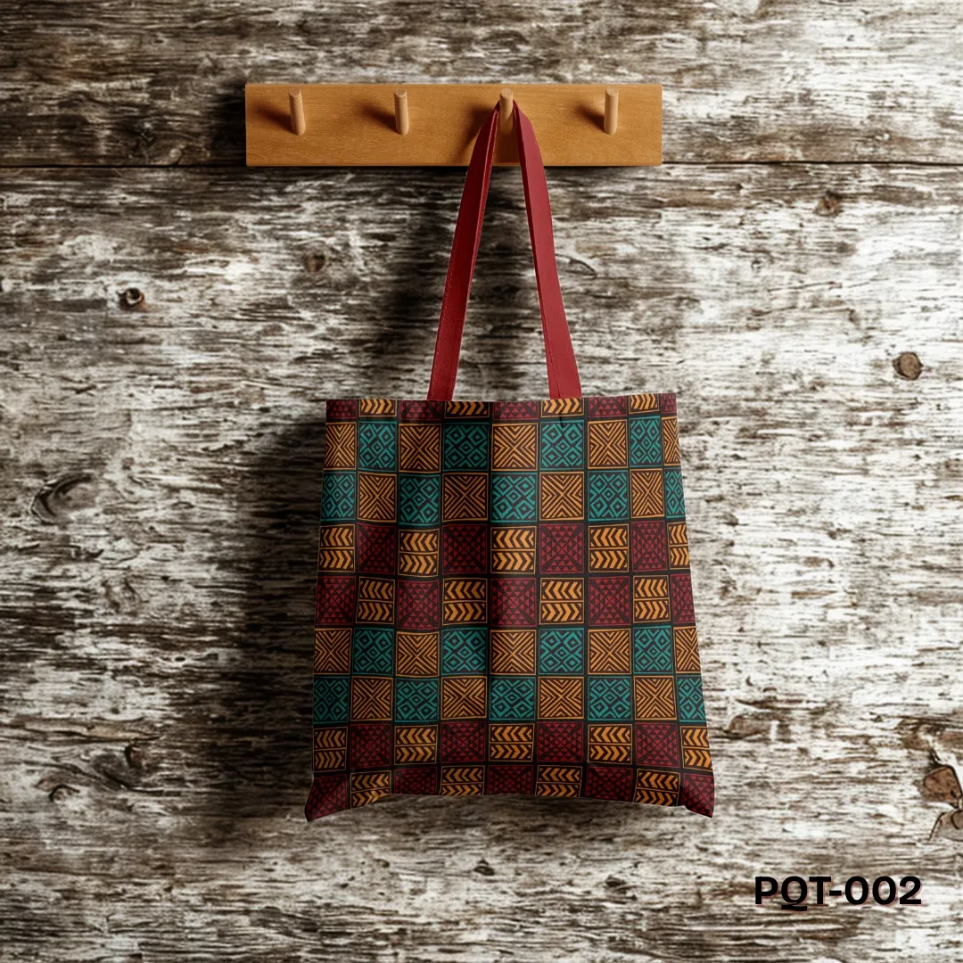 Ethnic Charm Tote Bag (PQT-002) featuring a repeating patchwork pattern of square tiles with tribal geometric symbols in teal, orange, and deep red, with red handles, hanging from a light wooden peg rack against a rustic weathered wooden plank wall.