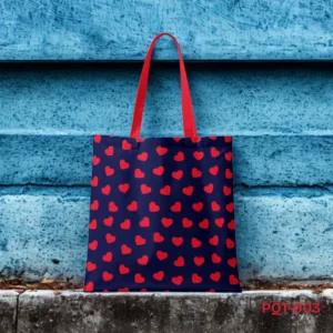 Navy Romance Tote Bag (PQT-003) featuring a repeating pattern of small red hearts on a navy blue background, with bright red handles, displayed upright on a concrete ledge against a textured blue painted wooden wall.