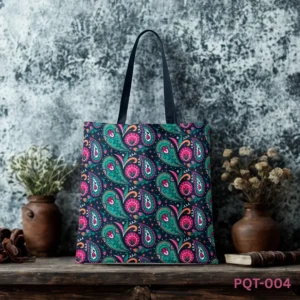CareCraft Paisley Tote Bag (PQT-004) featuring a repeating pattern of vibrant stylized paisley and floral motifs in teal, pink, and orange on a dark navy blue background, with black handles, displayed upright on a rustic wooden surface against a textured mottled grey wall with dried flower and book props.