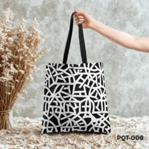 Noir Geometry Tote Bag (PQT-006) featuring a bold black and white abstract geometric line pattern, with black handles, held by a hand and displayed upright on a textured beige woven surface against a mottled grey concrete wall, with a vase of dried white flowers to the left.