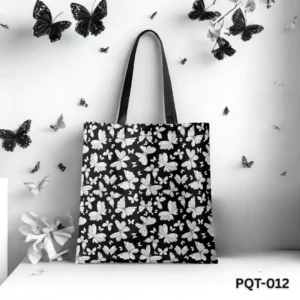 Black Wings Tote Bag (PQT-012) featuring a black background with a dense repeating pattern of white and grey stylized butterflies, with black handles, displayed upright on a white surface against a white wall decorated with black and white butterfly cutouts and a white flower.
