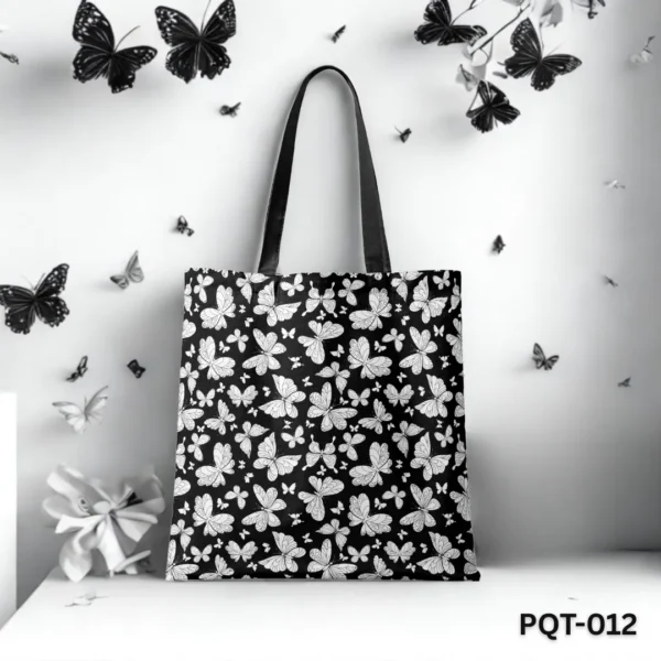 Black Wings Tote Bag (PQT-012) featuring a black background with a dense repeating pattern of white and grey stylized butterflies, with black handles, displayed upright on a white surface against a white wall decorated with black and white butterfly cutouts and a white flower.