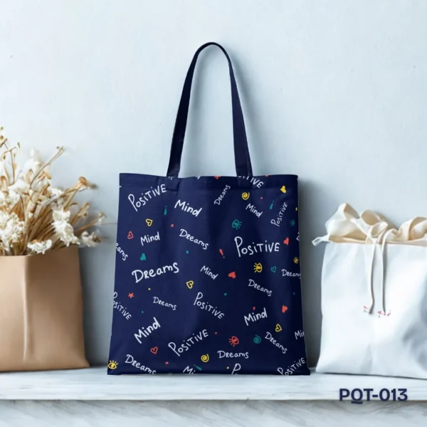 Posi Vibes Tote Bag (PQT-013), a navy blue canvas tote, featuring a repeating pattern of white handwritten-style words "Positive," "Mind," and "Dreams," with small colorful doodle accents, displayed upright on a white shelf against a light blue-grey wall with floral and bag props.