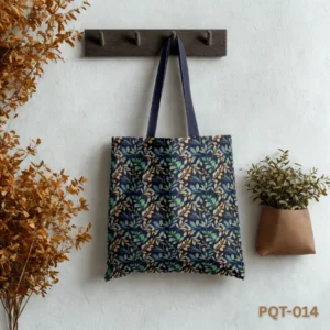 Amazonia Tote Bag (PQT-014) featuring a dense repeating pattern of green, blue-green, and yellow/orange leaves on a dark navy blue background, with matching navy blue handles, hanging from a dark wooden peg rack against a textured white wall, with dried orange-brown leafy branches to the left and a small potted green plant to the right.