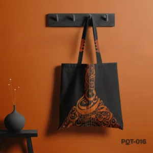 Raqs-e-harf Tote Bag (PQT-016), a black canvas tote, featuring an elegant orange/copper Arabic or Persian calligraphy design forming the shape of an Oud (musical instrument), with matching design accents on the black handles, hanging from a dark grey peg rack against a deep orange wall, with a dark grey vase and dried flowers on a small black table to the left.