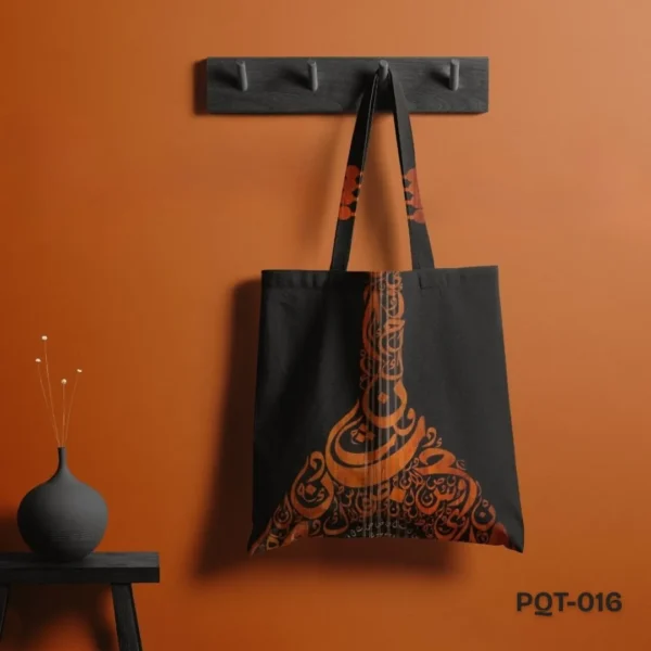 Raqs-e-harf Tote Bag (PQT-016), a black canvas tote, featuring an elegant orange/copper Arabic or Persian calligraphy design forming the shape of an Oud (musical instrument), with matching design accents on the black handles, hanging from a dark grey peg rack against a deep orange wall, with a dark grey vase and dried flowers on a small black table to the left.