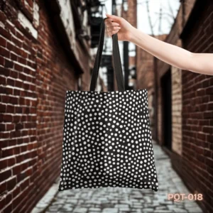 Classic Dots Tote Bag (PQT-018) featuring a black background with a dense pattern of white polka dots and black handles, held by a hand in a blurred urban alleyway with red brick walls and a cobblestone path.