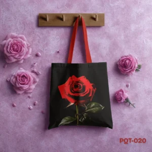 Red Rose Tote Bag (PQT-020), a black canvas tote, featuring a large, vibrant photorealistic print of a single red rose with green stem and leaves, with red handles, hanging from a light wooden peg rack against a textured purple-pink wall adorned with pink roses and petals.