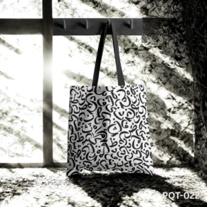 Callitote Tote Bag (PQT-022), a white canvas tote, featuring a dense repeating pattern of black Arabic or abstract calligraphic script, with black handles, hanging from a dark wooden peg rack in a dramatically lit, high-contrast black and white setting with strong shadows and light filtering through a window or foliage.