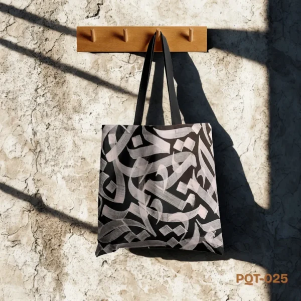 Golden Script Tote Bag (PQT-025), a black canvas tote, featuring a bold, abstract design of light pinkish-beige (or "golden" if it appears so) calligraphy-style brushstrokes, with black handles, hanging from a light wooden peg rack against a sunlit, textured beige concrete wall.