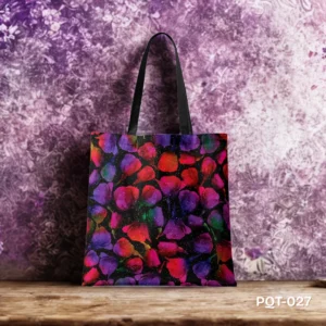 Galaxy Garden Tote Bag (PQT-027) featuring a vibrant, abstract design of overlapping stylized flower petal shapes in glowing jewel tones (purple, pink, red, blue, green) with white speckles resembling stars on a black background, with black handles, displayed upright on a rustic wooden surface against a textured purple and white wall.