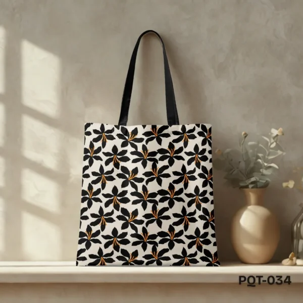 Savannah Blossom Tote Bag (PQT-034) featuring a repeating pattern of stylized black lily-like flowers with orange centers on a cream background, with black handles, displayed upright on a light-colored shelf against a sunlit beige textured wall with a beige vase of dried flowers to the right.