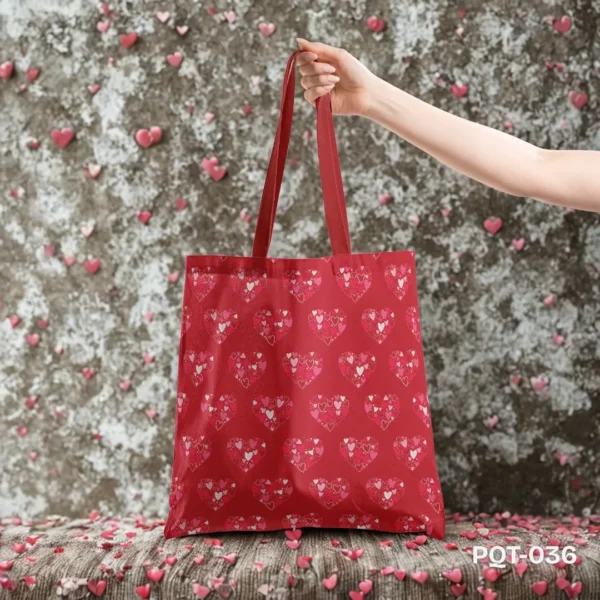 Scarlet Love Tote Bag (PQT-036), a vibrant red canvas tote, featuring a repeating pattern of large heart shapes filled with smaller pink and white hearts, with matching red handles, held by a hand and displayed upright on a textured surface with scattered pink hearts against a mottled grey wall also adorned with pink hearts.