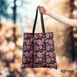 Sindhi Tradition Tote Bag (PQT-039) featuring a repeating pattern of intricate paisley and floral motifs in purple, pink, and yellow on a black background, with black handles, held by a hand against a blurred autumnal outdoor background with bokeh lights.