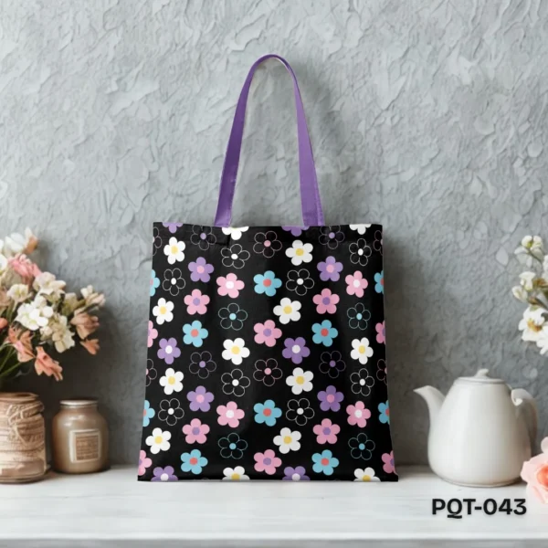 Starry Garden Tote Bag (PQT-043) featuring a repeating pattern of stylized pastel pink, purple, blue, and white simple flowers with yellow centers, and flower outlines, on a black background, with purple handles, displayed upright on a white surface against a textured grey wall with floral and teapot props.