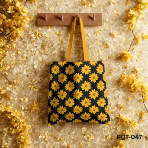 Sunkissed Tote Bag (PQT-047) featuring a repeating pattern of bright yellow sunflowers on a black background, with yellow handles, hanging from a light wooden peg rack against a textured beige wall adorned with scattered yellow petals and flower branches.