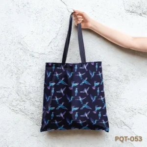 Azure Winged Voyage Tote Bag (PQT-053) featuring an all-over print of colorful hummingbirds in flight (teal, blue, purple) on a dark navy blue background, with matching navy handles, held by a hand against a textured white stucco wall.