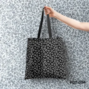 Midnight Circles Tote Bag (PQT-054) featuring an all-over print of hand-drawn white circles and dots of various sizes on a black background, with black handles, held by a hand against a black and white floral patterned wallpaper.