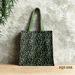 Herbal Muse Tote Bag (PQT-058) featuring an all-over print of light green slender leaves on a black background, with light green handles, displayed upright on a light wooden surface against a mottled grey concrete wall, with green foliage in the upper left corner.