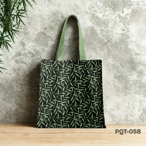 Herbal Muse Tote Bag (PQT-058) featuring an all-over print of light green slender leaves on a black background, with light green handles, displayed upright on a light wooden surface against a mottled grey concrete wall, with green foliage in the upper left corner.