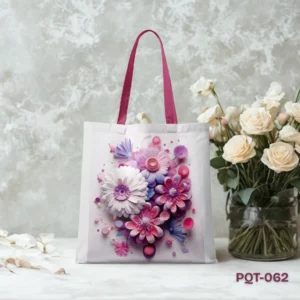 Violet Garden Tote Bag (PQT-062), a white canvas tote, featuring a vibrant 3D-style illustrative bouquet of pink, purple, and white layered paper-cut flowers, with magenta/pink handles, displayed upright on a white marble surface against a textured light grey wall with a vase of white roses to the right and scattered petals.
