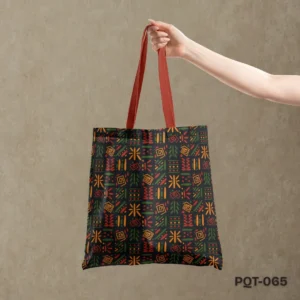 Artisan Carry Tote Bag (PQT-065) featuring a repeating pattern of abstract tribal-inspired geometric symbols in orange, green, and red on a dark grey or black background, with contrasting orange-red handles, held by a hand against a textured muted brown wall.
