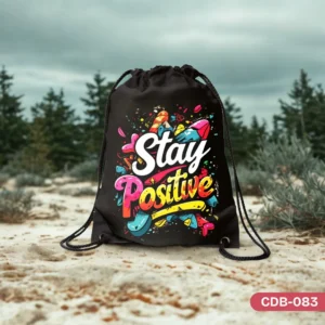 "Vibrant Vibe Drawstring Bag (CDB-083) featuring a colorful graffiti-style 'Stay Positive' design, photographed outdoors on a sandy surface with blurred trees in the background."