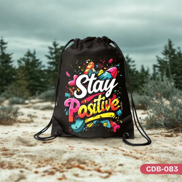 "Vibrant Vibe Drawstring Bag (CDB-083) featuring a colorful graffiti-style 'Stay Positive' design, photographed outdoors on a sandy surface with blurred trees in the background."