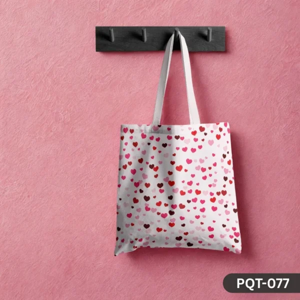 Love Drizzle Tote Bag (PQT-077) featuring a white background with an all-over print of scattered small hearts in various shades of red and pink, with white handles, hanging from a dark wooden peg rack against a textured pink wall.