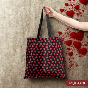 Velvet Valentine Tote Bag (PQT-078), a black canvas tote, featuring a repeating pattern of small, textured red glitter-like hearts, with black handles, held by a hand against a textured beige burlap background with scattered red velvet hearts.