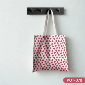 Blush Beats Tote Bag (PQT-079), a white canvas tote, featuring a repeating pattern of small, textured red glitter-like hearts, with light pink or white handles, hanging from a dark grey peg rack against a plain white wall.