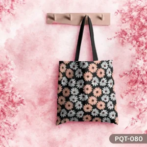 Wildflower Whimsy Tote Bag (PQT-080) featuring a black background with a repeating pattern of stylized peach and light blue/white daisies, with black handles, hanging from a light wooden peg rack against a textured pink watercolor-style wall with cherry blossom branches framing the sides.