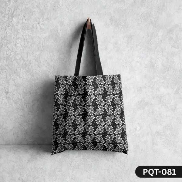 Botanic Noir Tote Bag (PQT-081) featuring a dense repeating pattern of stylized white and grey flowers on a black background, with black handles, hanging from a single light-colored peg against a textured white wall with a subtle floral imprint.