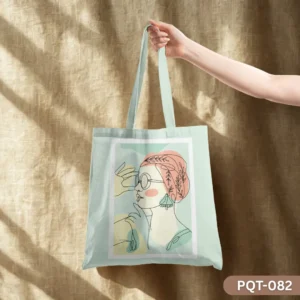 Headwrap Harmony Tote Bag (PQT-082), a light mint green canvas tote, featuring a minimalist line art illustration of a woman in profile wearing a peach headwrap, round glasses, and a teal earring, with abstract pastel color block accents, held by a hand against a textured beige burlap wall with dappled sunlight.