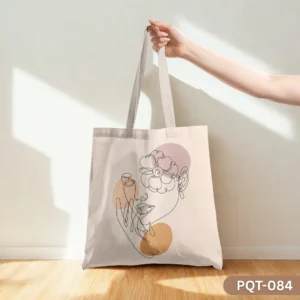 Soft Statement Tote Bag (PQT-084), a light beige canvas tote, featuring a black continuous line art illustration of a woman's face with a flower over her eye, accented by muted pink and tan circular color blocks, held by a hand and resting on a light wooden floor against a sunlit beige wall.
