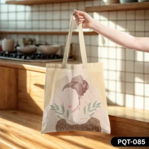The Calm Carry Tote Bag (PQT-085), a light beige canvas tote, featuring a soft watercolor-style illustration of a woman's profile with a floral headpiece and botanical elements in muted earthy tones, held by a hand and resting on a wooden kitchen counter with a sunlit tiled backsplash.