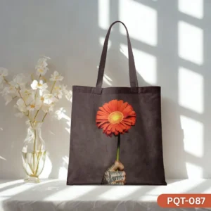 Botanik Noir Tote Bag (PQT-087), a dark brown canvas tote, featuring a photorealistic print of a tattooed hand holding a vibrant orange gerbera daisy, displayed upright on a white surface against a sunlit white wall with a glass vase of white flowers to the left.
