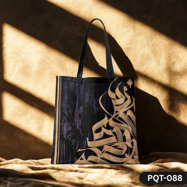 CalligraVibe Tote Bag (PQT-088) featuring a dark textured background with elegant golden Arabic or abstract calligraphy art, and black handles, displayed upright on a draped beige fabric against a sunlit earthy brown textured wall.