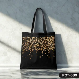 Golden Cascade Tote Bag (PQT-089), a black canvas tote, featuring an elegant design of cascading golden Arabic or abstract calligraphy with scattered gold square accents, displayed upright on a polished light surface against a sunlit textured white marble wall.