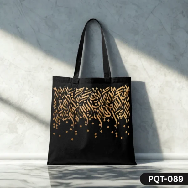 Golden Cascade Tote Bag (PQT-089), a black canvas tote, featuring an elegant design of cascading golden Arabic or abstract calligraphy with scattered gold square accents, displayed upright on a polished light surface against a sunlit textured white marble wall.