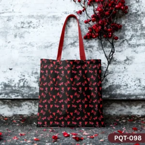 Luna Rose Tote Bag (PQT-098) featuring a black background with a repeating pattern of small red hearts, leaves, and floral elements, with red handles, displayed upright on a dark textured surface with scattered red petals, against a distressed white brick wall with a branch of red berries.