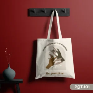 The Positiva Tote Bag (PQT-101), a white canvas tote, featuring an arched design with two hands gently holding a botanical element with butterfly accents, and the inspirational quotes "GOOD VIBES ONLY FOR YOURSELF" and "Be positive," hanging from a dark grey peg rack against a deep red wall, with a teal vase on a small black table to the left.