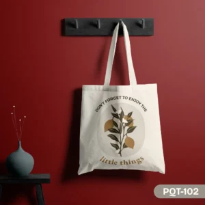The Olive Verse Tote Bag (PQT-102), a white canvas tote, featuring a central circular design of a lemon-like branch with yellow fruit and white flowers, framed by the inspirational quote "DON'T FORGET TO ENJOY THE little things," hanging from a dark grey peg rack against a deep red wall, with a teal vase on a small black table to the left.