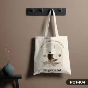 Serenity Voyage Tote Bag (PQT-104), a white canvas tote, displaying a mystical design with hands, a crescent moon, stars, and botanical elements, framed by the quotes "WE'LL GO THERE, FAST OR SLOW." and "Be grateful," hanging from a dark grey peg rack against a muted brown wall, with a teal vase and dried flowers on a small black table to the left.