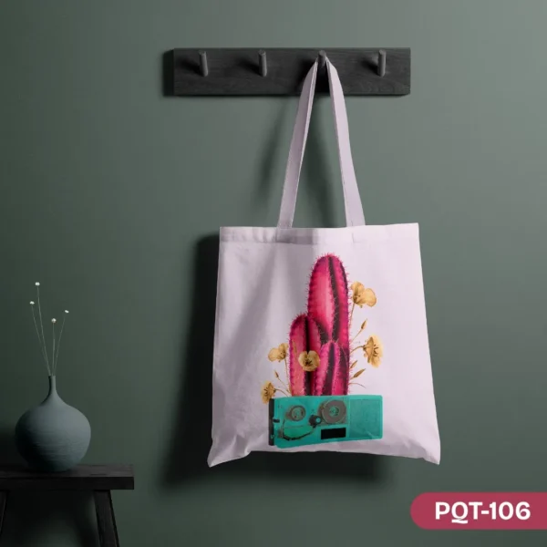 Cactus Groove Tote Bag (PQT-106), a white canvas tote, featuring a vibrant pink cactus with yellow flowers emerging from a retro teal radio/cassette player design, hanging from a dark grey peg rack against a dark teal wall, with a teal vase and dried flowers on a small black table to the left.
