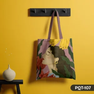 Dreamy Petals Tote Bag (PQT-107) featuring an illustrative design of a woman's serene face in profile, surrounded by large red and yellow flowers and green leaves, on a dusty pink background, hanging from a dark grey peg rack against a vibrant yellow wall, with a beige vase and dried flowers on a small black table to the left.