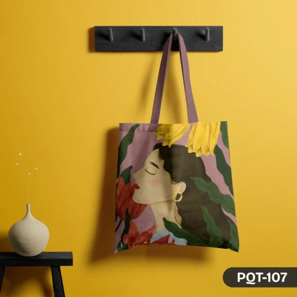 Dreamy Petals Tote Bag (PQT-107) featuring an illustrative design of a woman's serene face in profile, surrounded by large red and yellow flowers and green leaves, on a dusty pink background, hanging from a dark grey peg rack against a vibrant yellow wall, with a beige vase and dried flowers on a small black table to the left.
