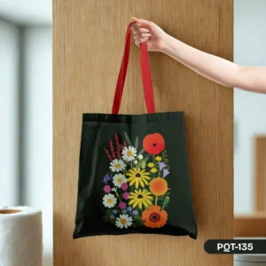 Hand holding the Wildflower Waltz Tote Bag (PQT-135) featuring a vibrant print of various wildflowers on a dark background with bold red straps, against a light wood panel