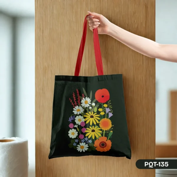 Hand holding the Wildflower Waltz Tote Bag (PQT-135) featuring a vibrant print of various wildflowers on a dark background with bold red straps, against a light wood panel
