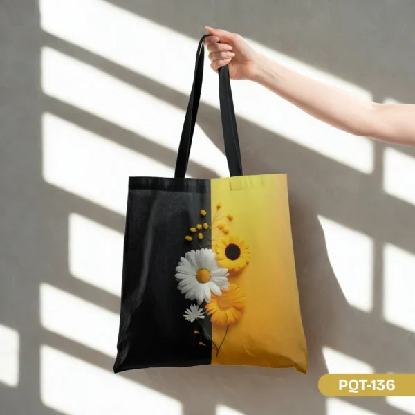 Hand holding the Daybreak Noir Tote Bag (PQT-136) featuring a striking split black and yellow design with sunflower and daisy florals, black straps, against a light grey wall with window shadows.